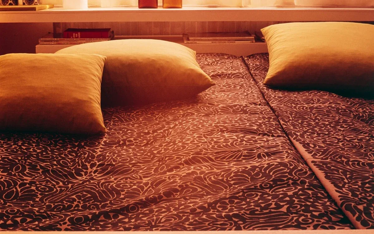 A patterned bedspread and cushions sit below a wooden ledge beside a sunlit window