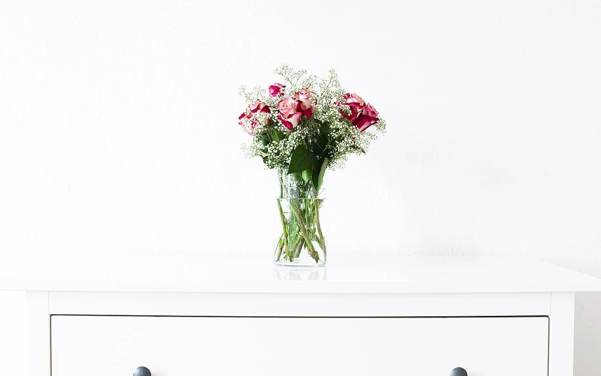 A vase of pink and pale flowers rests on a pale drawer unit against a light wall