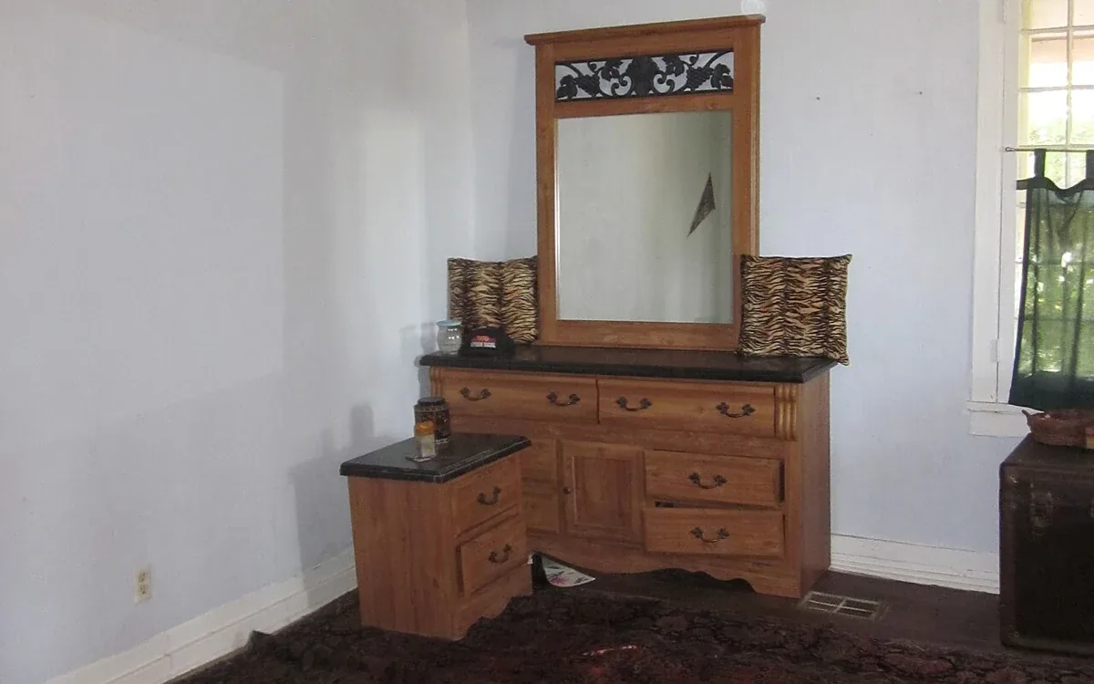 A wooden dresser and mirror stand against a pale wall in a room with little other furniture