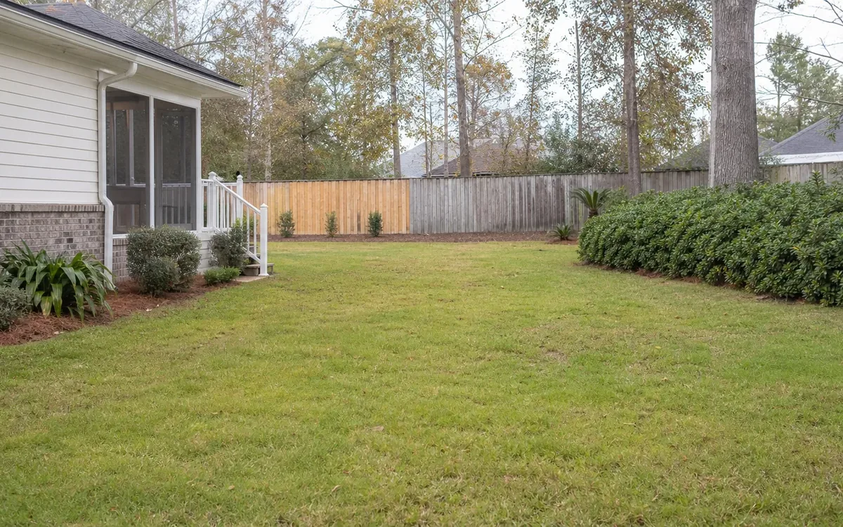 An open grassy backyard beside a screened porch, with shrubs, trees and a wooden fence bordering the clear lawn.