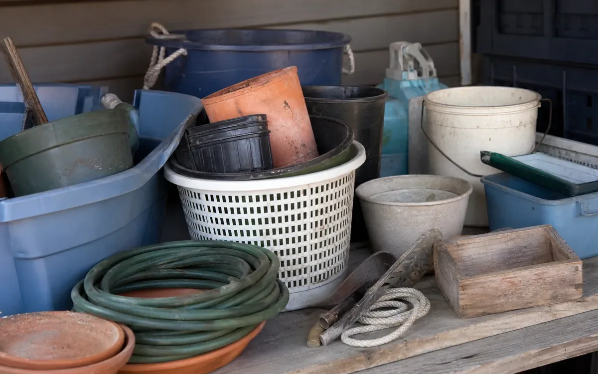 Pots, buckets, plastic bins, a coiled garden hose, rope, and a wooden box crowd shelves against an exterior wall.