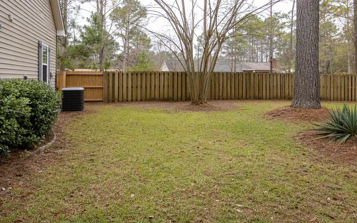 Fenced backyard with patchy grass, trees, shrubs, and an outdoor air conditioning unit beside a beige-sided house.