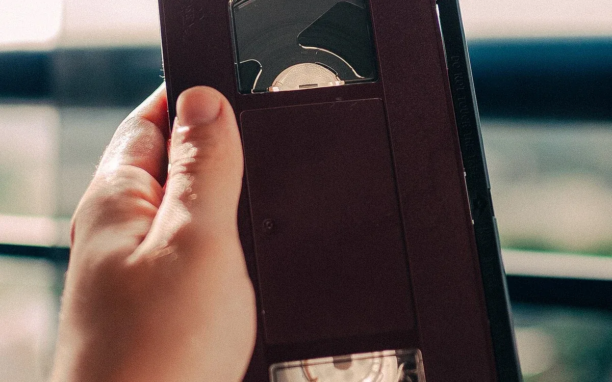 A hand holds an unlabelled black videotape near a bright window