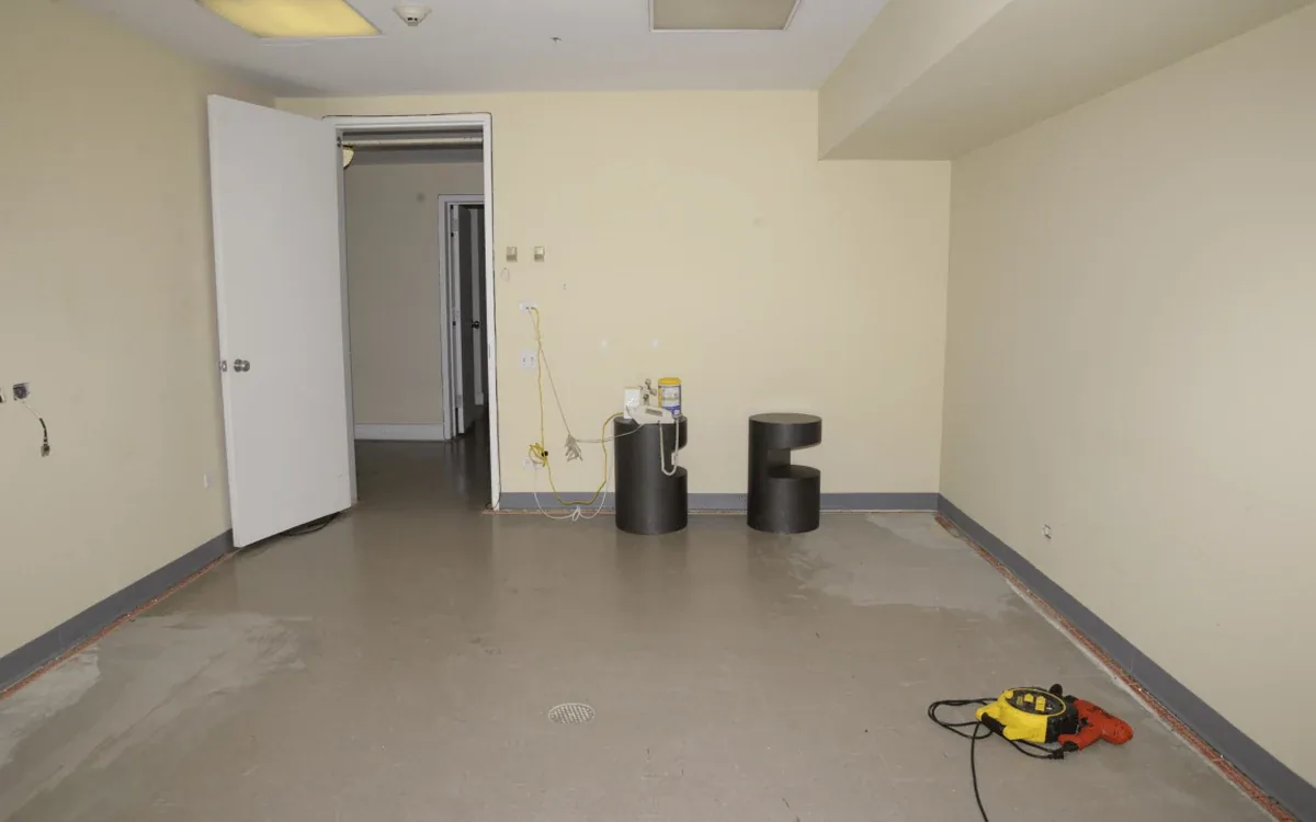 An empty pale-walled room has a stripped floor, an open doorway and a small floor tool near the wall