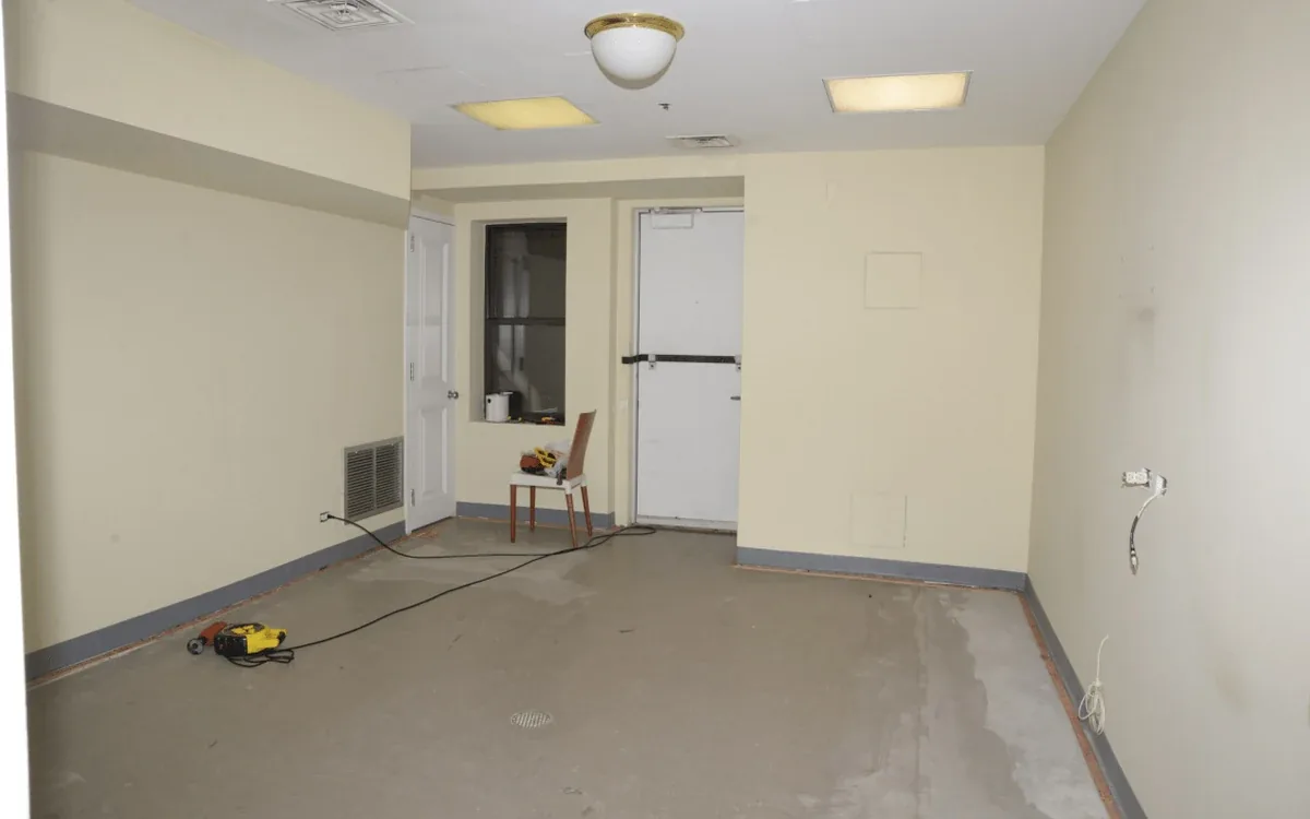 A room with a stripped floor opens toward a kitchen and doorway, with a coiled cable lying along the floor