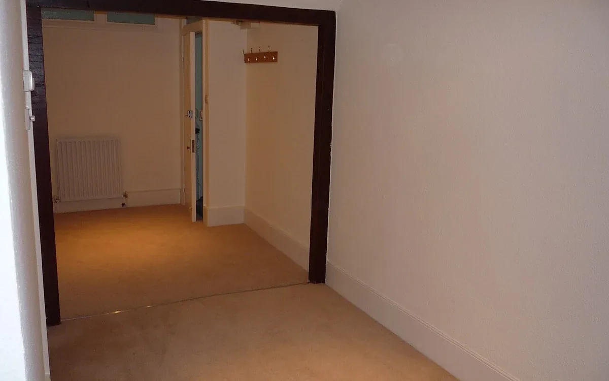 Two empty pale-walled rooms are connected by a broad doorway with dark wooden trim and a carpeted floor