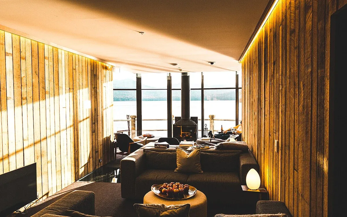 A long wood-paneled room contains grouped chairs, low tables and lamps beside tall sunlit windows