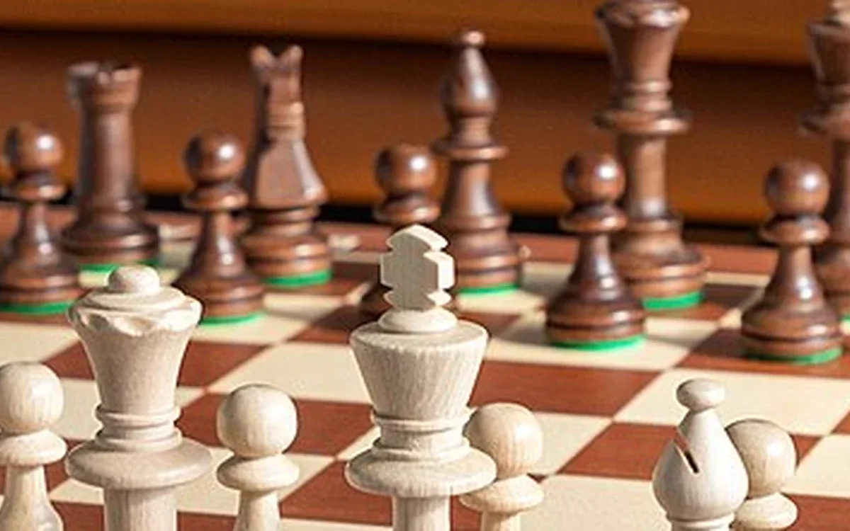 Light and dark wooden chess pieces stand on brown and cream squares of a board beside a tan sofa