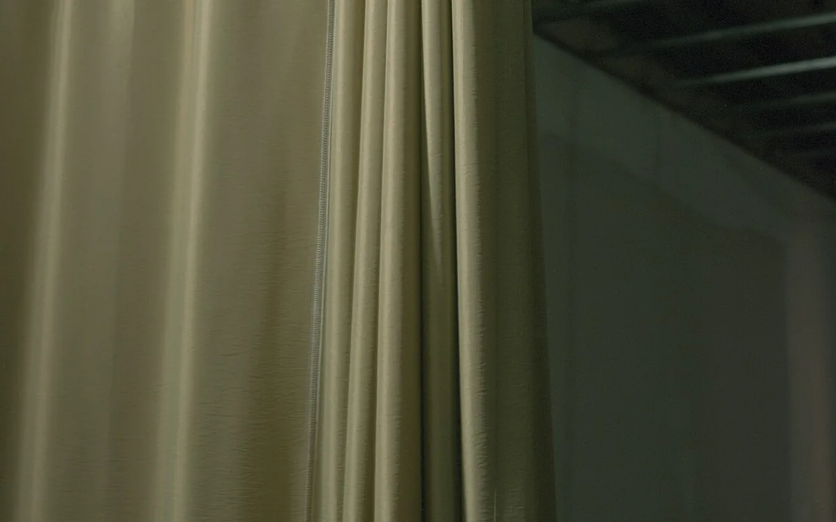 Close-up of plain beige curtains hanging in vertical folds beside a dark interior.