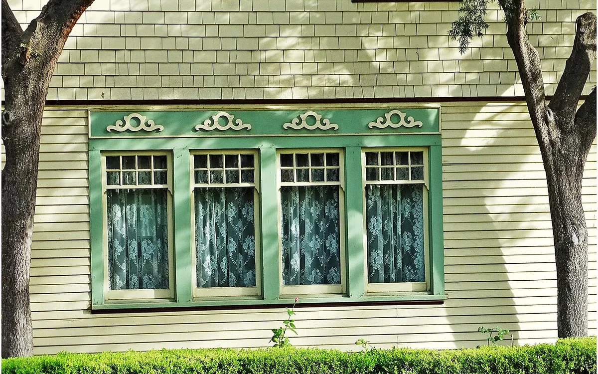 Lace curtains in four green-trimmed windows on a house with pale wooden siding.