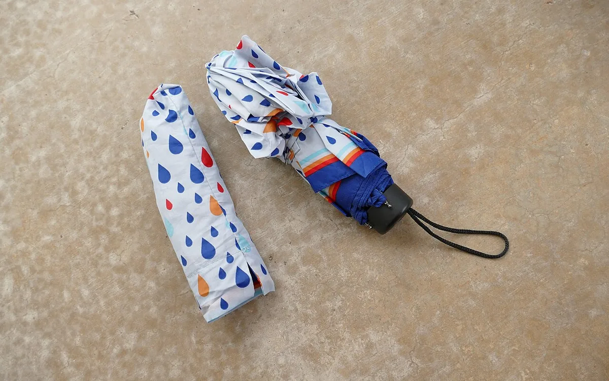 A compact umbrella with a multicolored raindrop pattern and black wrist loop lies beside its matching sleeve on a beige surface.