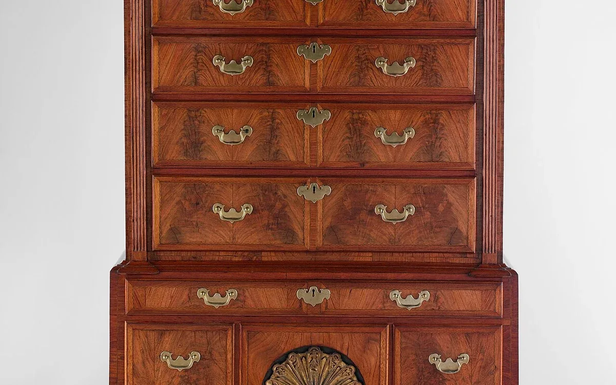 A tall wooden chest of drawers with curved legs and a decorative top stands against a plain light background
