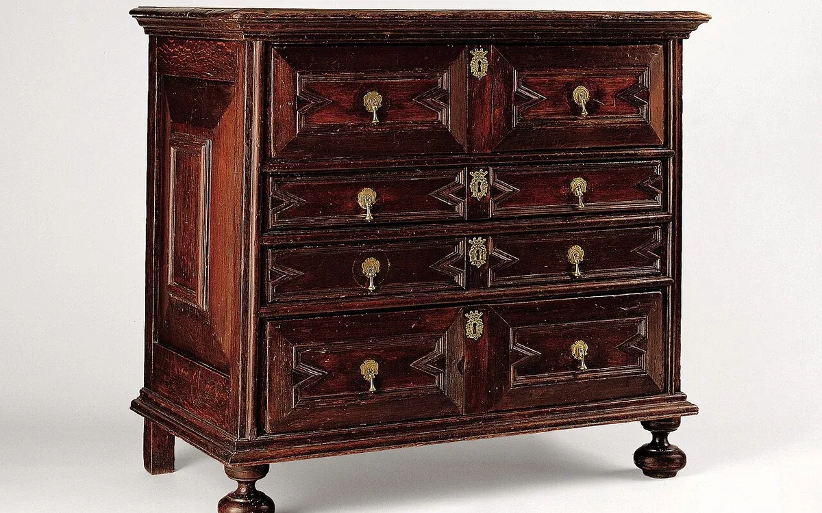 A dark wooden antique chest with six drawers and brass handles stands alone against a pale background