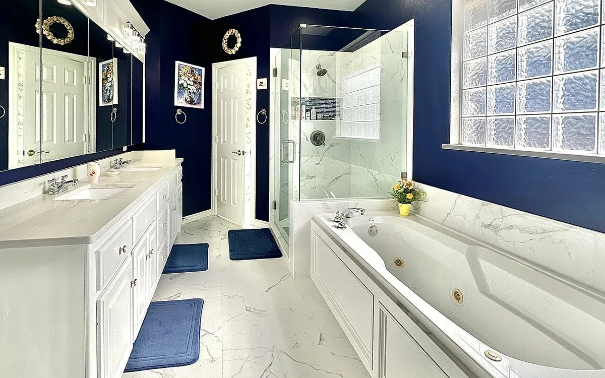 A bathroom has white drawers and cupboards, a double vanity, a bath and a glass shower against dark blue walls
