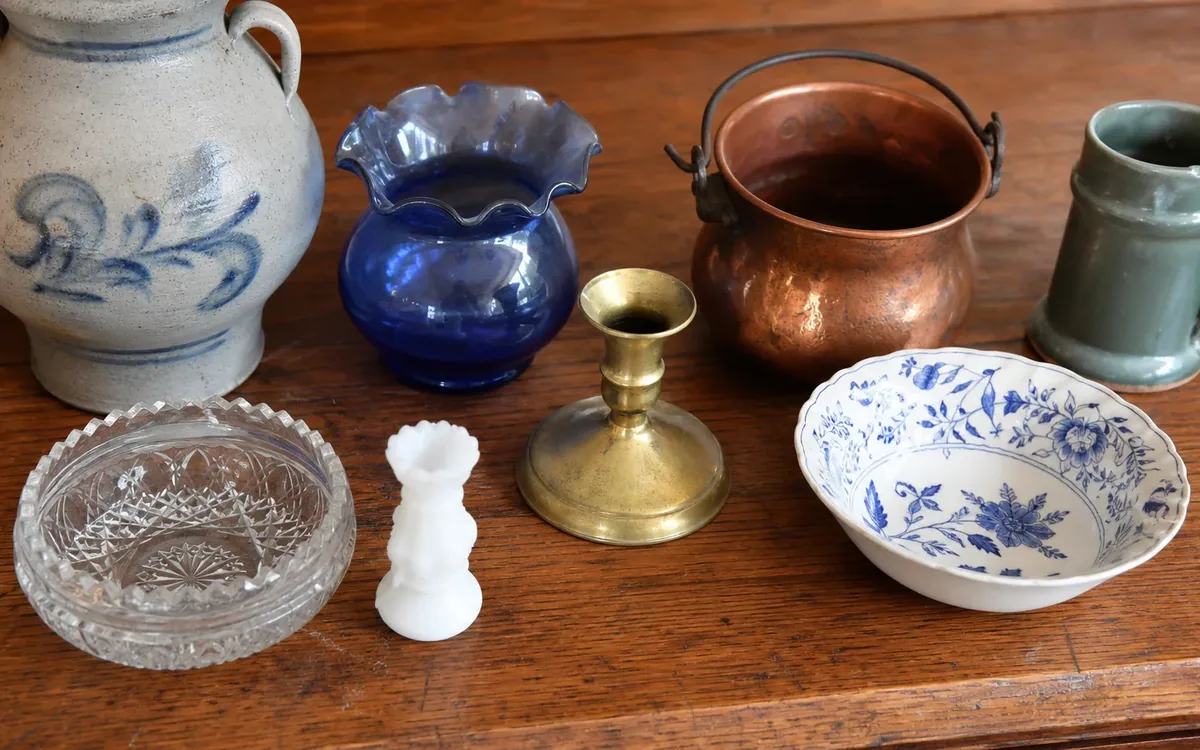 Eight ceramic, glass, and metal household objects arranged on a wooden table, including a patterned bowl and brass candlestick.