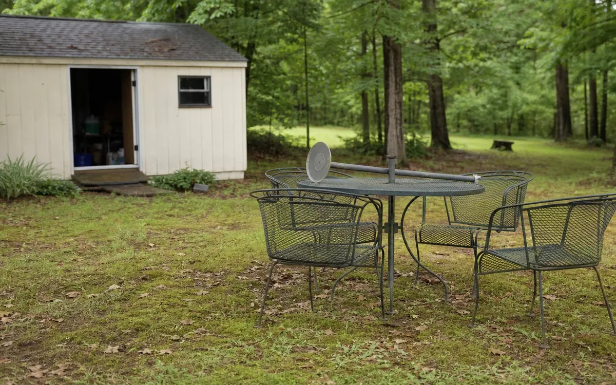 A metal patio table and three mesh chairs sit on patchy grass beside a light-colored shed with an open doorway.