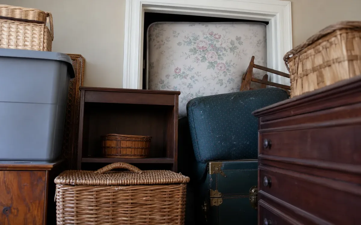 A doorway is blocked by an upright floral mattress, a tilted blue chair, trunks, furniture, and wicker baskets.