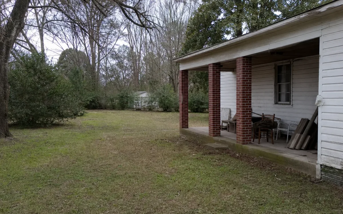 A white-sided house has a covered porch with brick columns, chairs, and leaning boards beside a grassy yard and trees.
