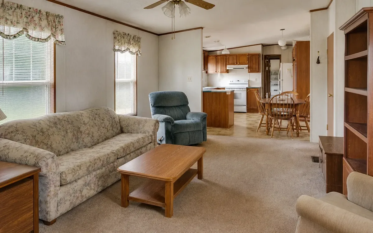 Furnished mobile home interior with sofa, recliner, tables, shelving, dining area, and kitchen.