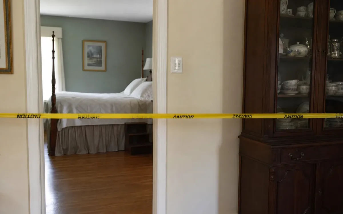 Yellow caution tape blocks access to a furnished bedroom beside a china cabinet filled with dishes.