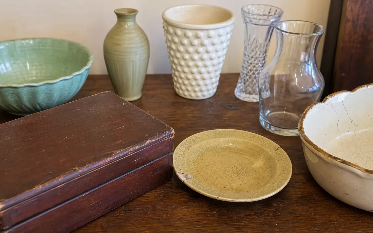 A scuffed wooden box, chipped yellow plate, and visibly cracked cream bowl sit on a wooden table with vases and a green bowl.