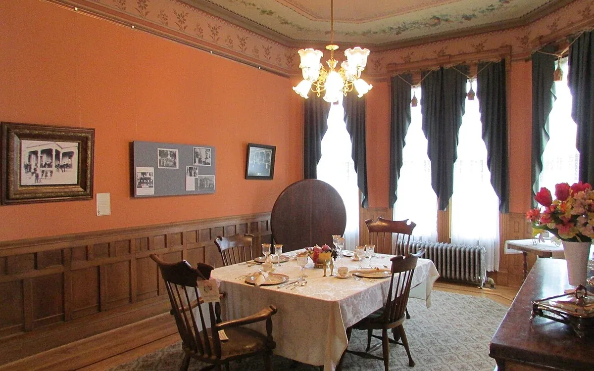 A wooden dining table and chairs stand in an orange-walled room beside tall windows with dark curtains