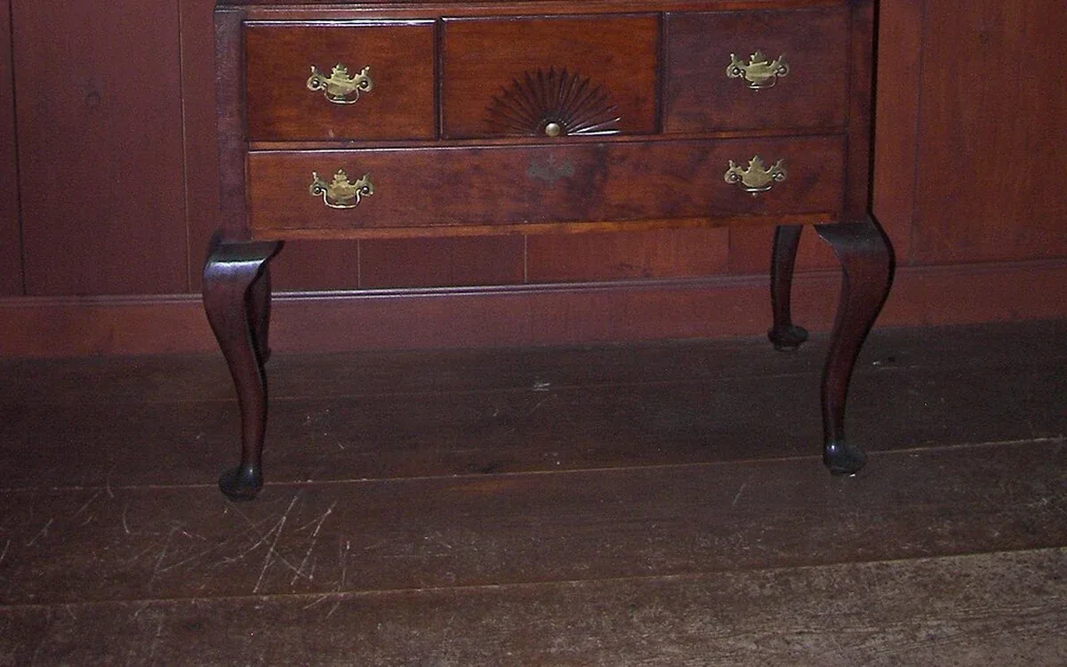 The lower drawers and curved feet of a wooden chest stand against a dark wall