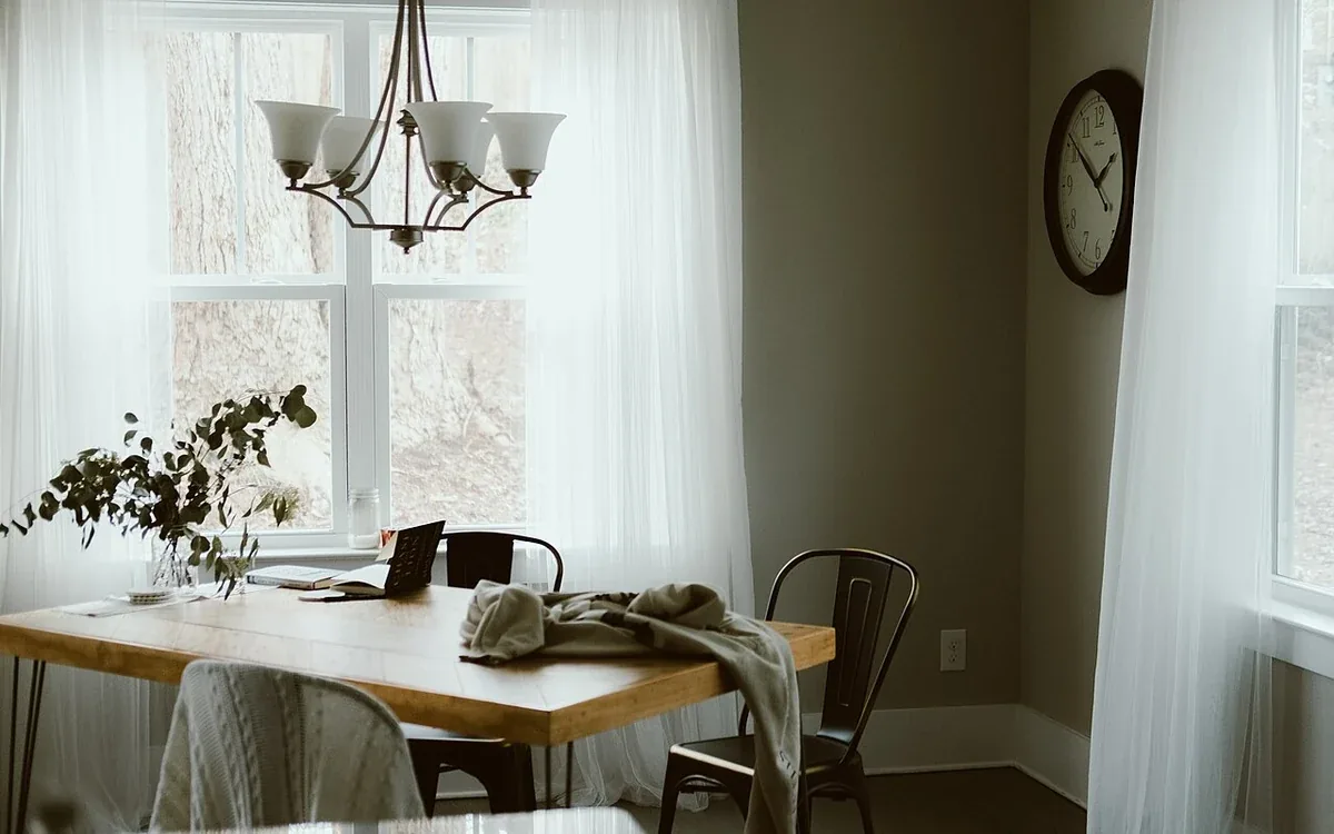 A dining table with chairs and a loose cloth stands beside broad curtained windows