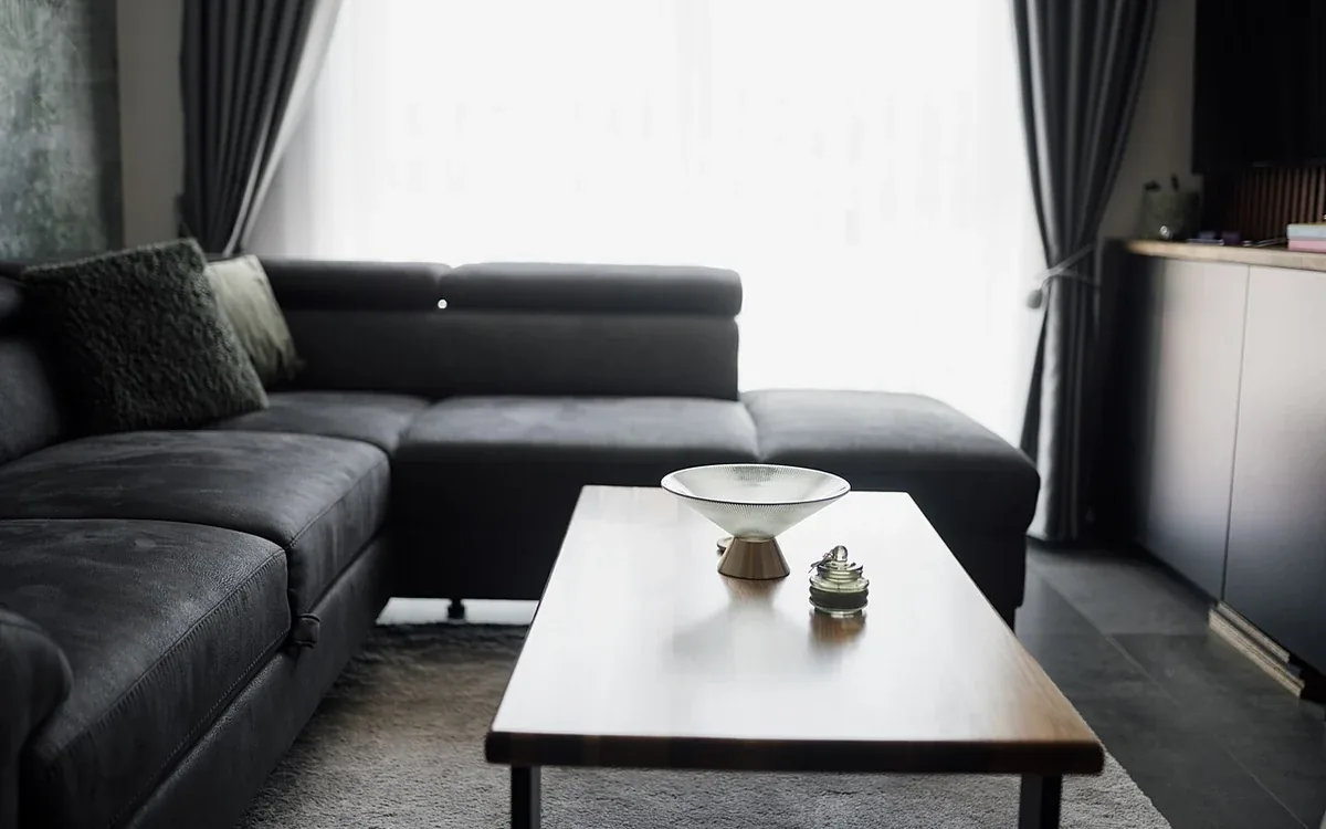 A dark sectional sofa sits beside a low pale table near a bright curtained window