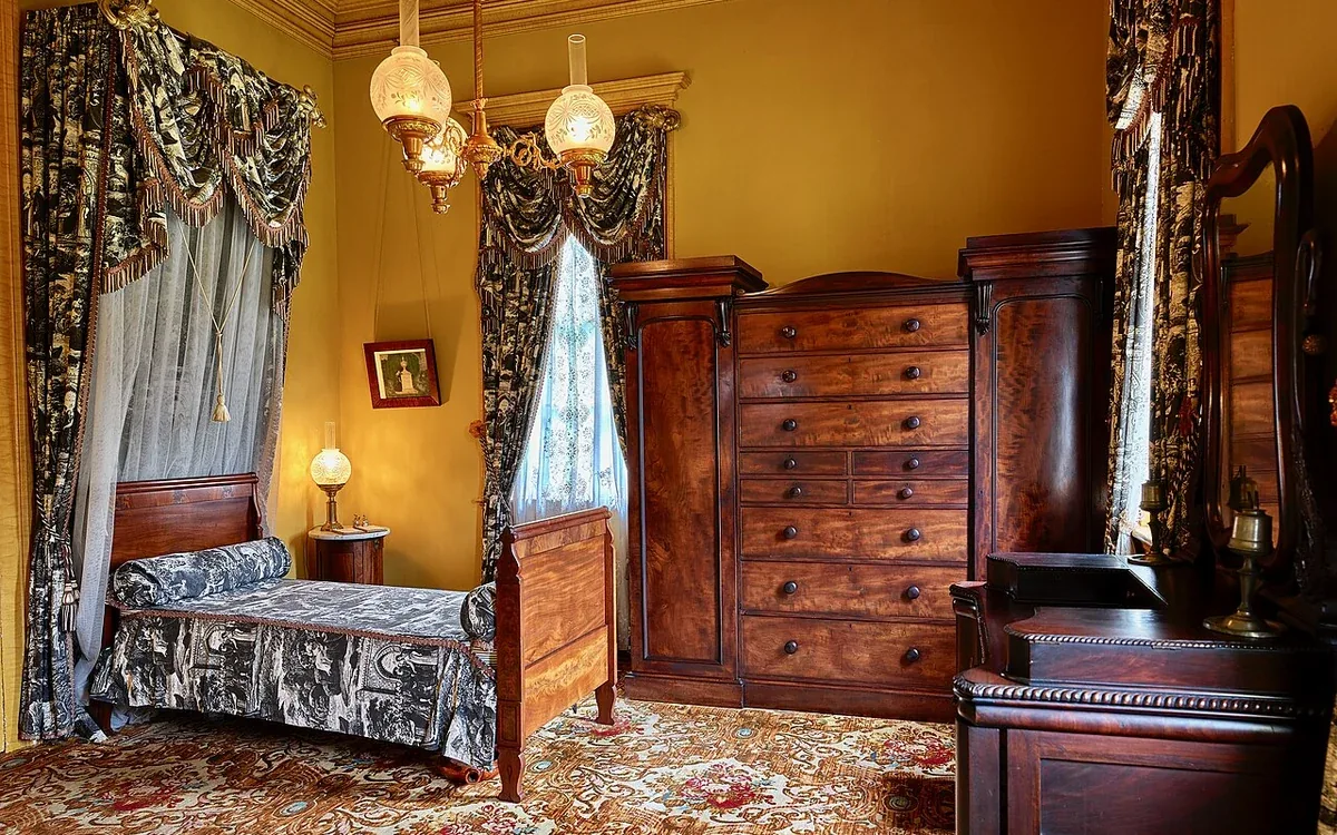 A bed and tall wooden chest stand among patterned curtains and other bedroom furniture