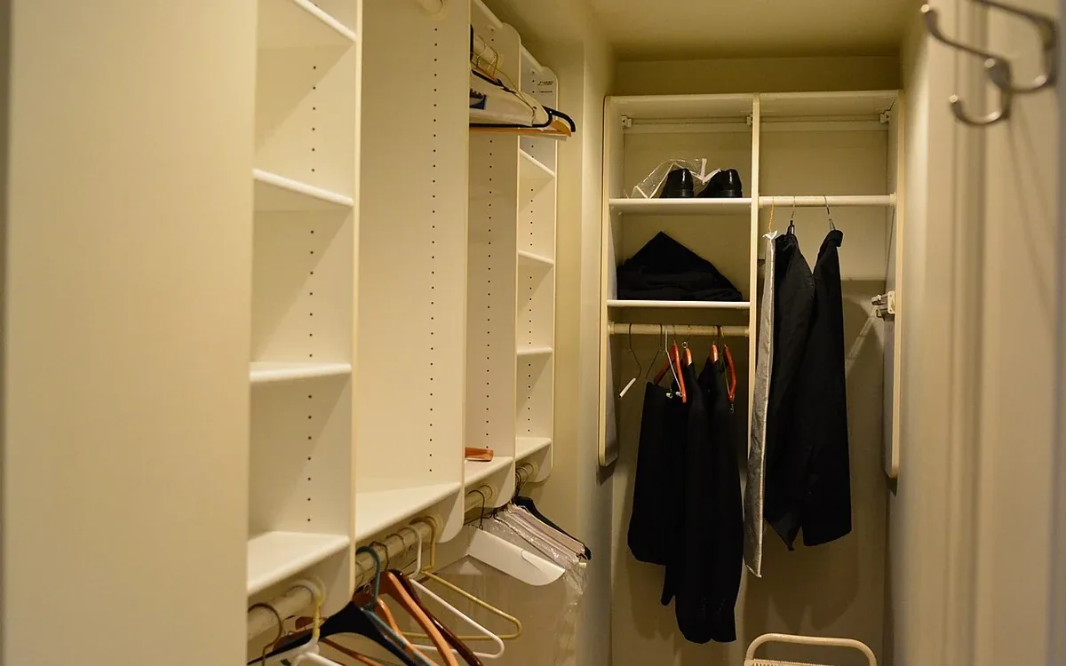 An open closet contains pale shelves, hanging clothes, empty hangers and a small chair