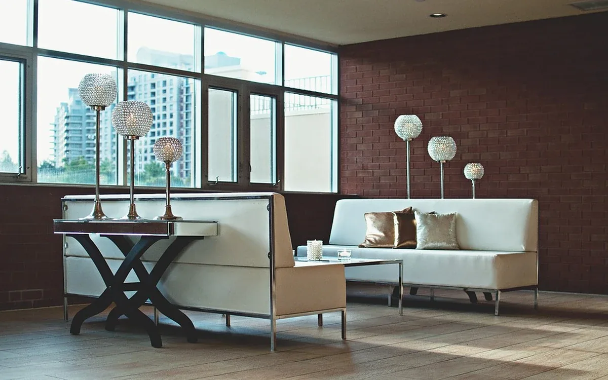 Pale upholstered seats and a narrow dark table stand beside broad windows and a brick wall