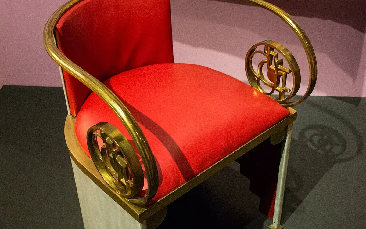 A red upholstered armchair with decorative metal arms and legs stands against a pale wall