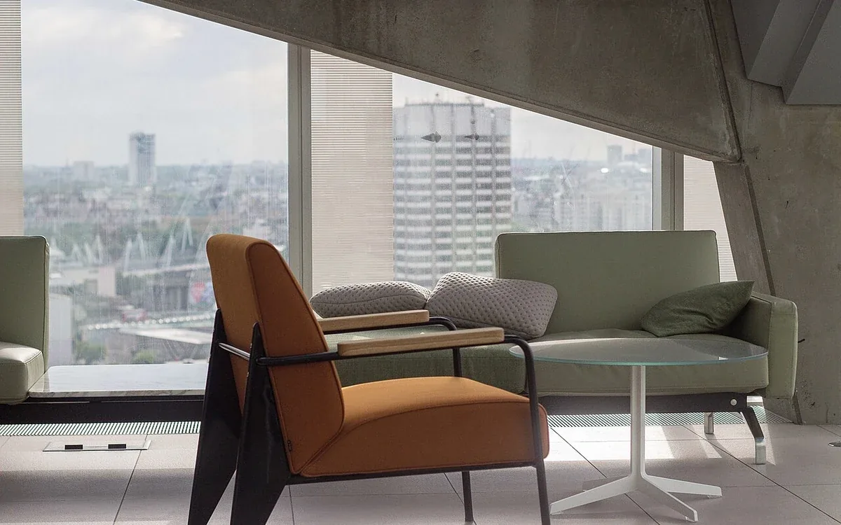 An armchair and a pale sofa stand beside broad windows overlooking buildings