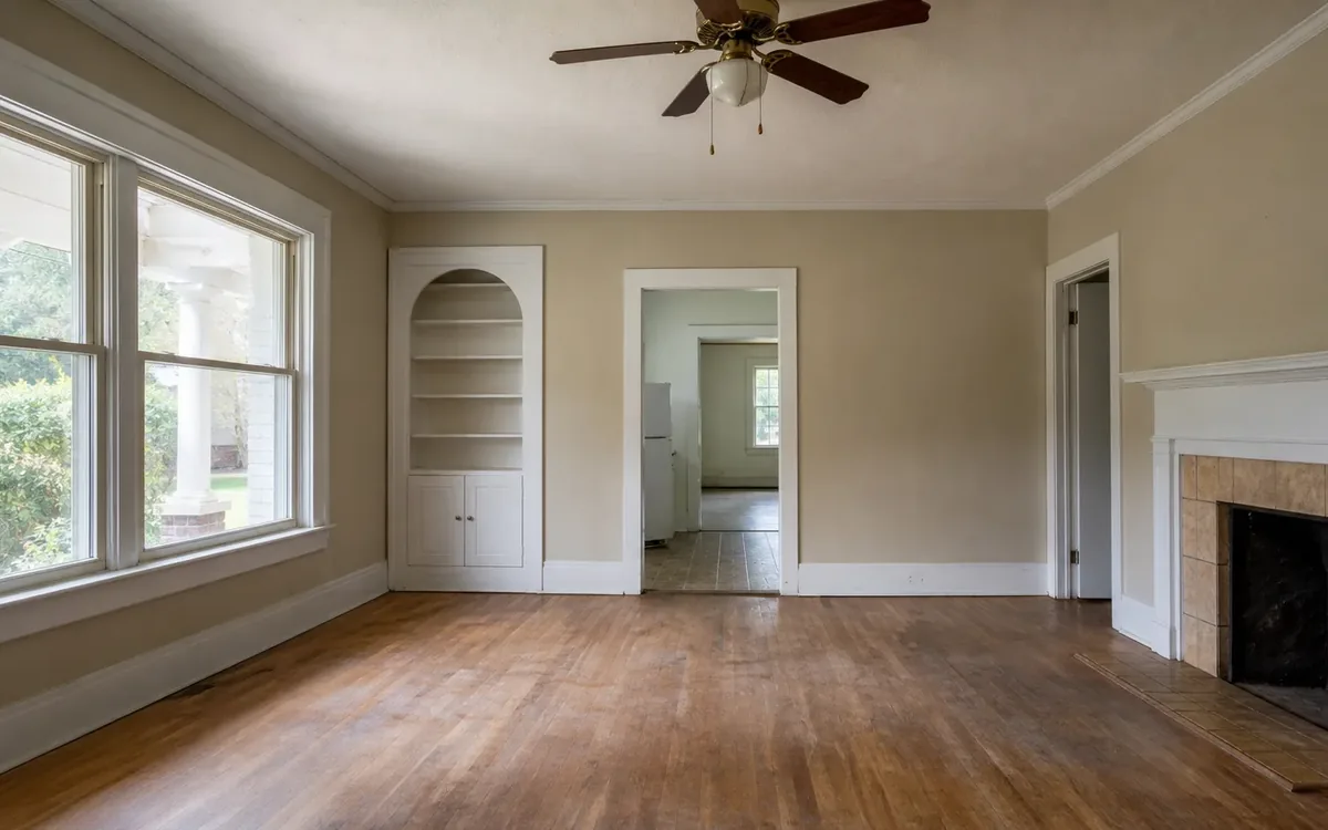 Empty room with worn hardwood flooring, large windows, built-in shelves, a ceiling fan, and a tiled fireplace.