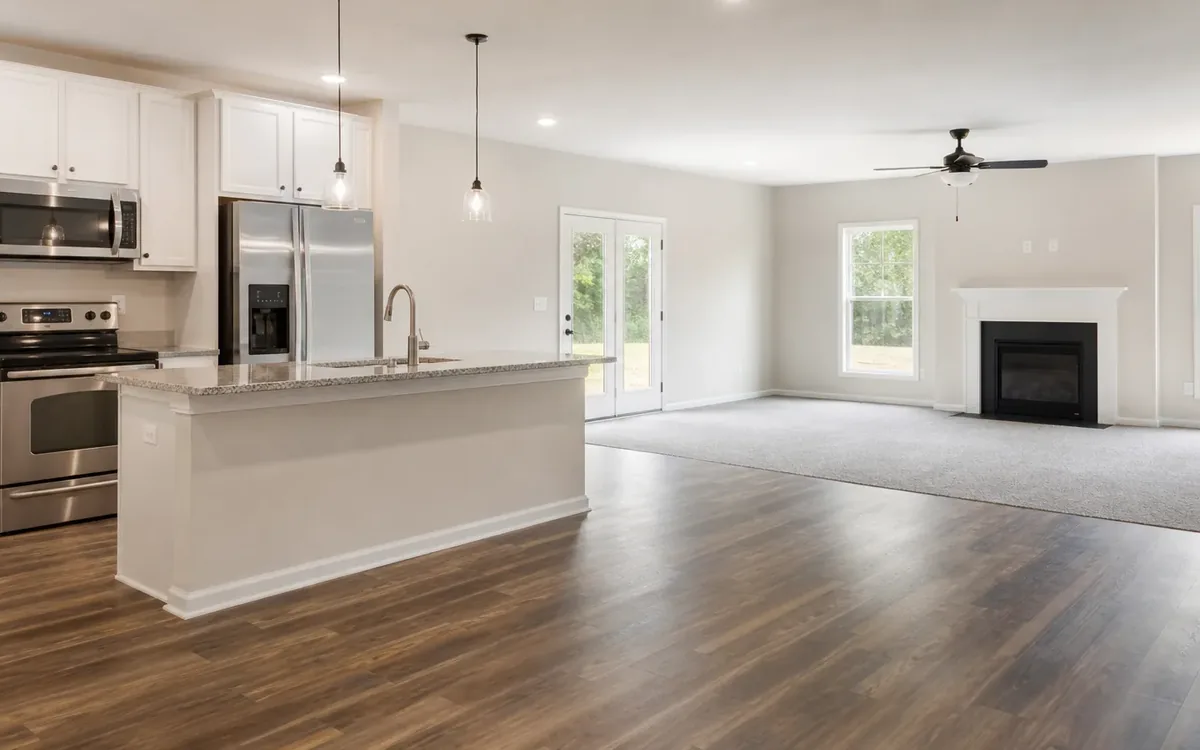 Empty open-plan kitchen and living room with white cabinets, stainless steel appliances, wood flooring, carpet, and a fireplace.