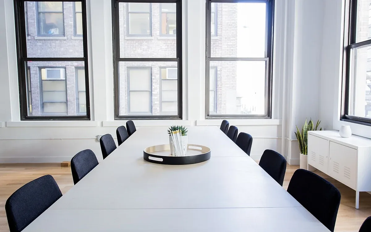A long clear white table with dark chairs stands beside tall windows and a radiator