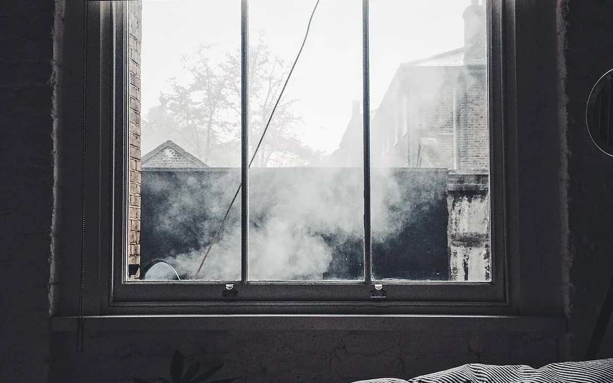 Rumpled bedding lies beneath a window partly covered by a dark roller shade, with daylight along the edges