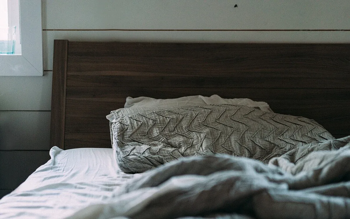 Light pillows and rumpled covers rest against a dark wooden headboard beside a pale paneled wall