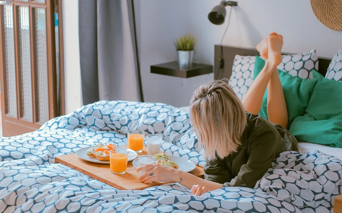 A woman rests on a patterned bed with her legs raised beside a breakfast tray, pillows and a wall lamp