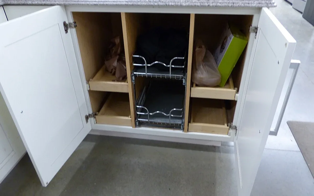 Two white cupboard doors stand open below a counter, exposing wooden dividers and a metal pull-out rack