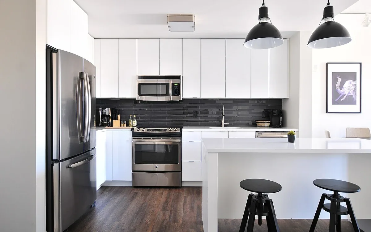 A white kitchen has an island, dark stools, stainless appliances and black pendant lights above the counter