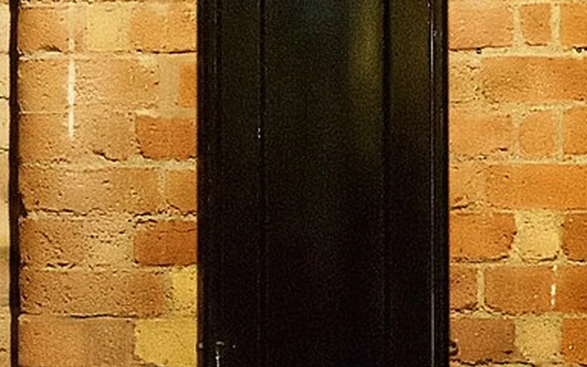 The dark wooden lower cabinet of a tall clock stands against a warm brick wall, with its narrow door closed