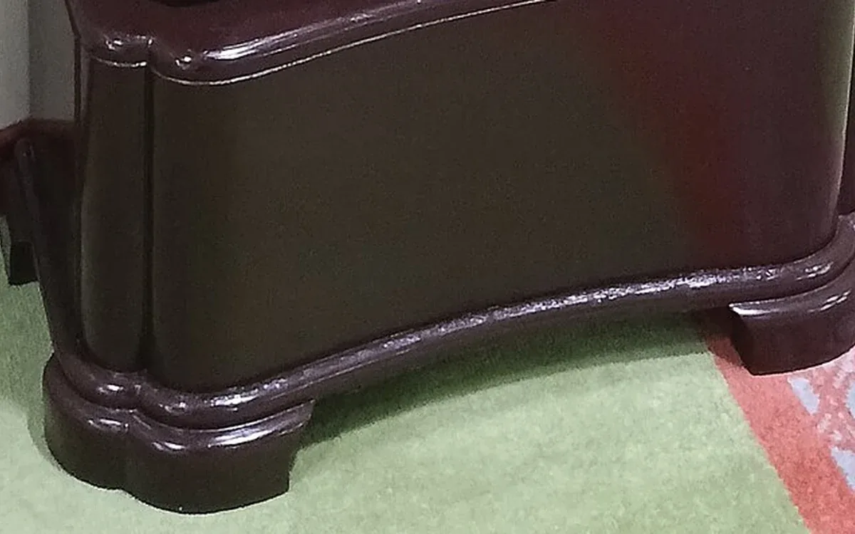 A curved dark wooden clock base stands on a light floor beside pale walls and a wooden skirting board