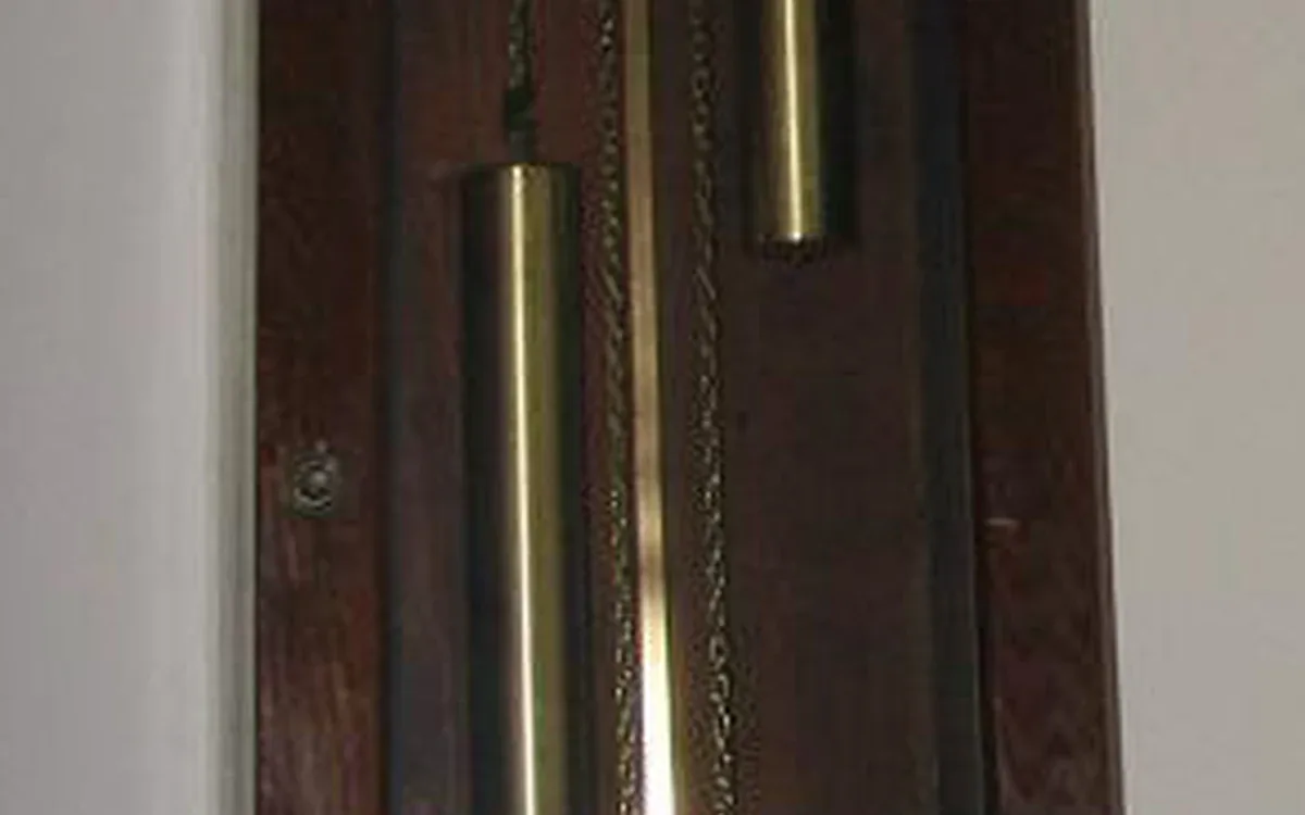Brass-colored cylinders and chains are visible behind the glass door of a dark wooden tall-clock case beside a pale wall