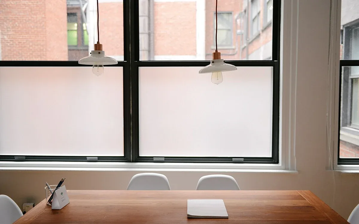 A wooden table with a chair and a small pale notebook stands beneath a broad window