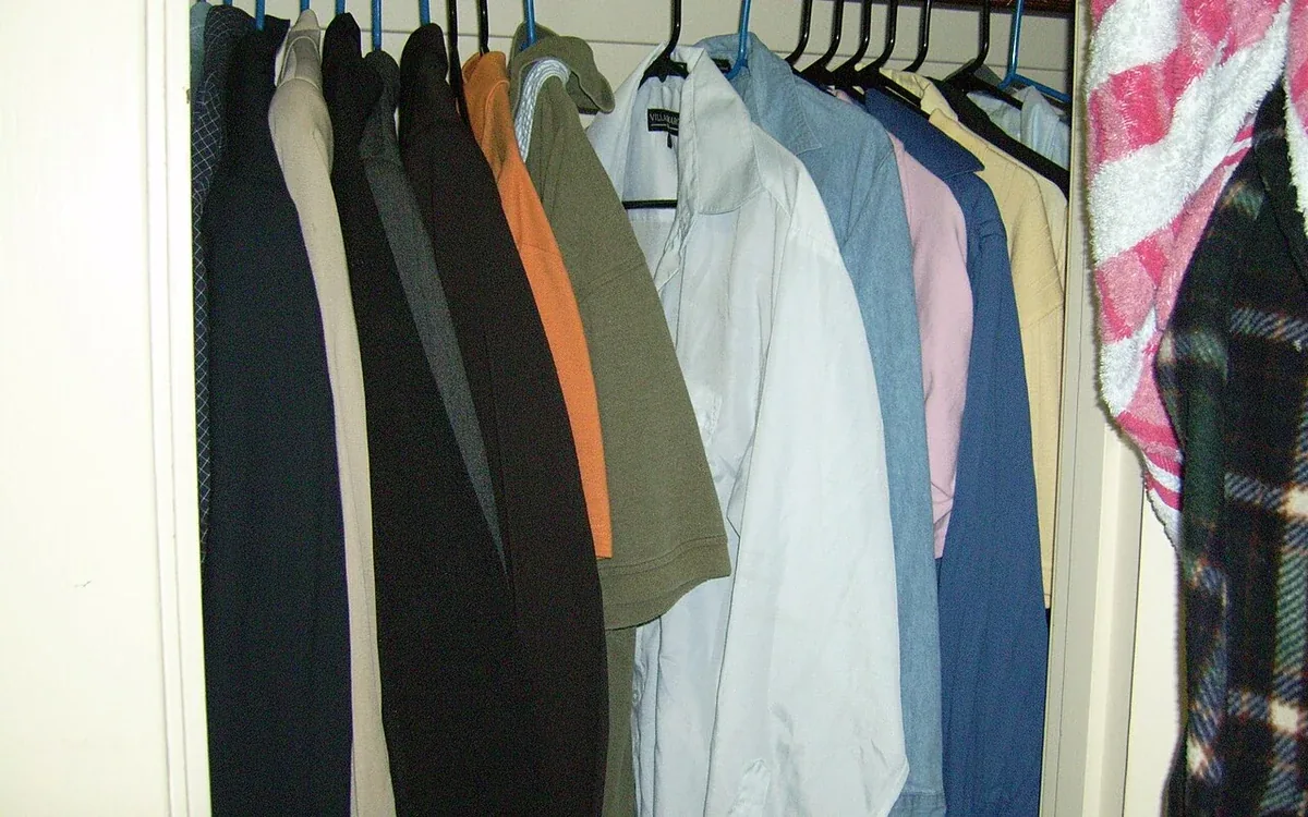 Coats and shirts in light, dark and warm colors hang close together on a rail inside a shallow closet