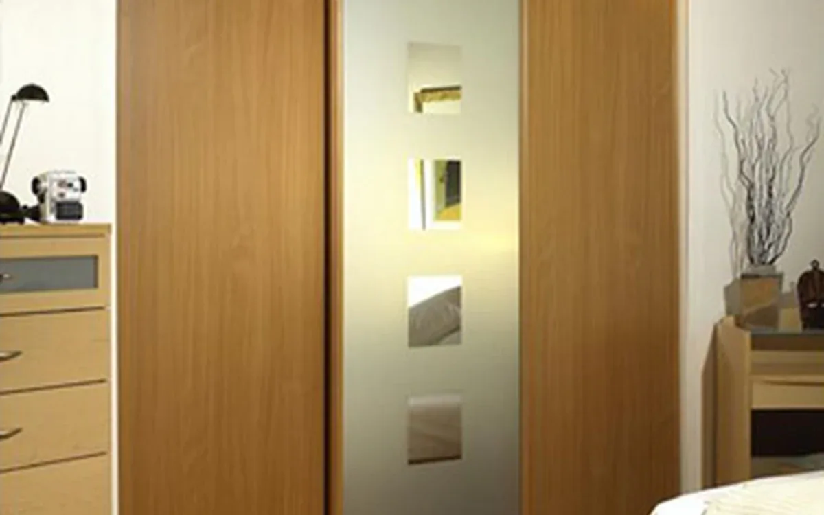 A wooden sliding wardrobe with a frosted-glass center panel stands beside the edge of a bed and drawers