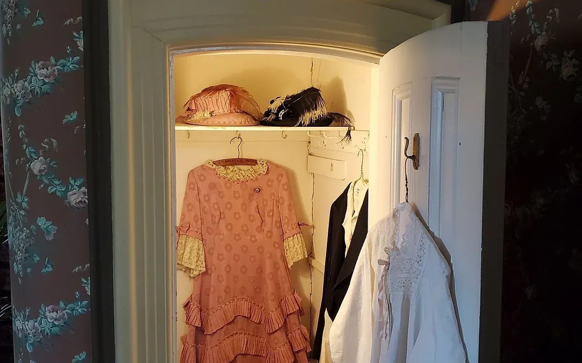Light and pink clothing hang inside a small open closet set into a blue patterned wall beside other belongings