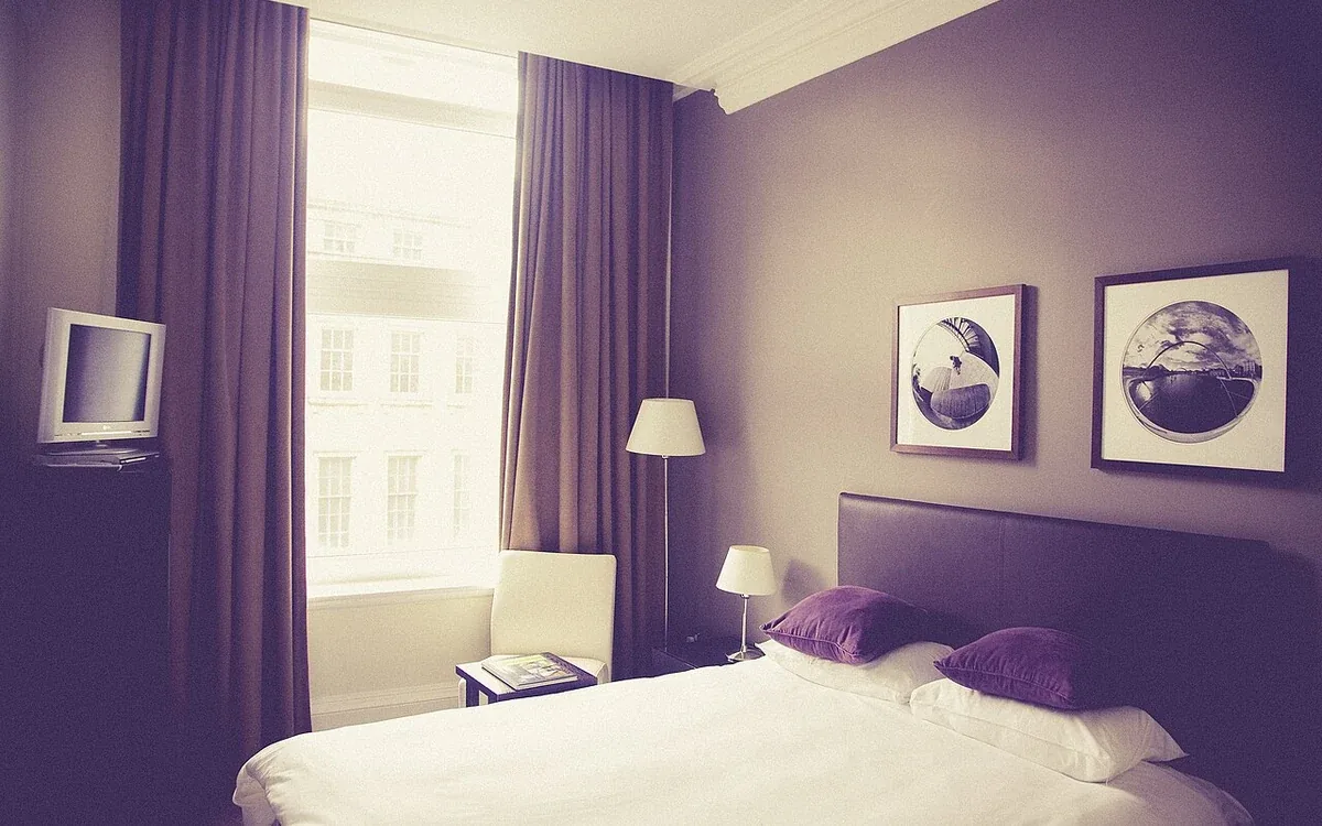 A bed with pale bedding stands beside a window with purple curtains, beneath framed pictures on a purple wall
